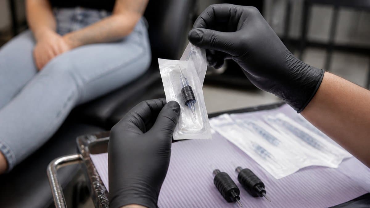 Close-up of a tattoo artist's gloved hands peeling open a sealed, single-use needle cartridge in front of a seated client in a clean, brightly lit studio