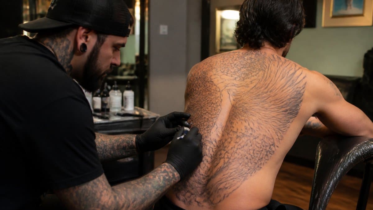 Professional tattoo artist applying large-scale design across client's back, showing how the tattoo follows natural shoulder blade contours and spine alignment in well-lit studio