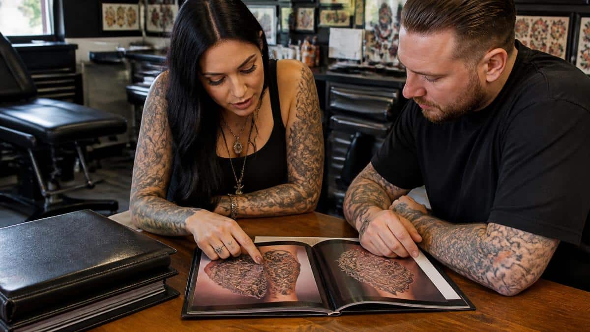 A tattoo artist and a client reviewing a printed portfolio together at a clean, well-lit consultation table inside a modern tattoo studio
