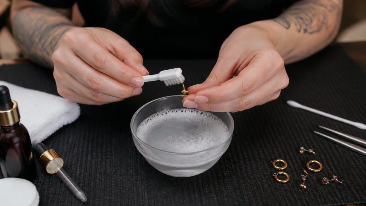 Close-up of hands gently cleaning earring posts with a soft brush and mild soapy water in a small bowl with natural lighting