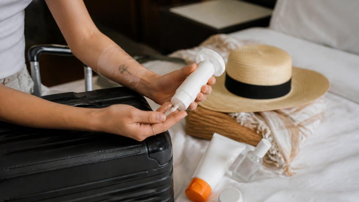 Close-up of a traveler's arm with a small fresh tattoo wrapped in protective film, resting on a suitcase while they apply fragrance-free lotion from a small travel bottle, with sunscreen and a wide-brim hat nearby on a hotel bed
